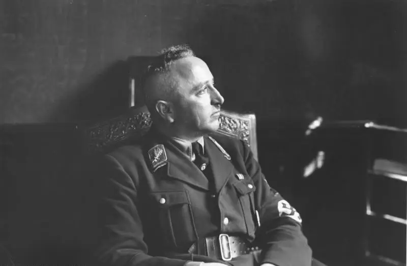 A man in a dark party uniform with an armband sits in a carved chair, looking to one side