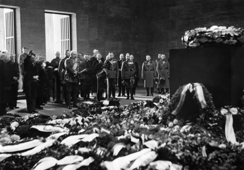 Officers and men in dark coats stand in a high stone hall beside a dark block bearing a wreath, the floor covered with wreaths and ribbons