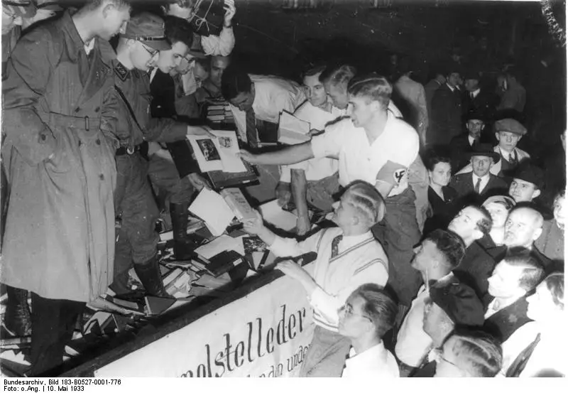 Young men, several in uniform, pass books down from a lorry hung with a banner while a crowd looks on at night