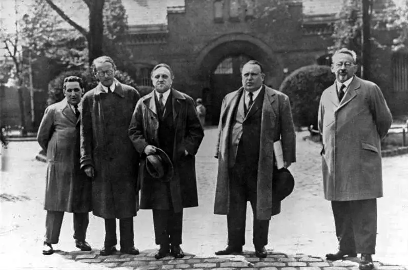 Five men in long coats standing in a row on cobblestones in front of a brick gatehouse.