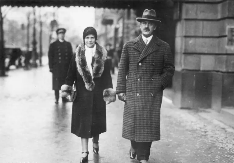 A man in a checked overcoat and hat walks beside a woman in a fur-collared coat along a wet pavement; a uniformed doorman stands behind.