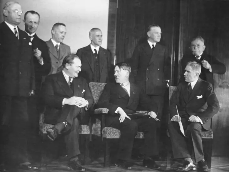 Group photograph of nine men in dark suits, three seated in armchairs and six standing behind them.