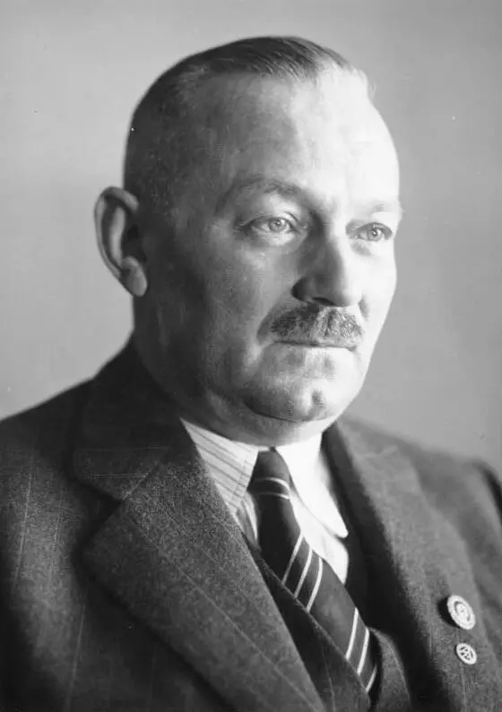 Portrait of a heavy-set man with close-cropped hair and a moustache, wearing a pinstriped suit, striped tie and a lapel badge.