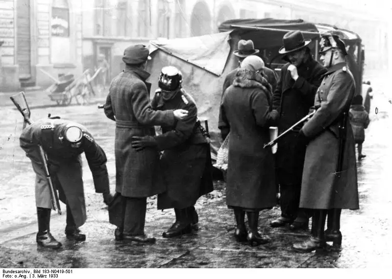 Uniformed police in shako helmets, some holding rifles, frisk men in overcoats and hats on a wet street beside a canvas-covered lorry, while a woman stands by.