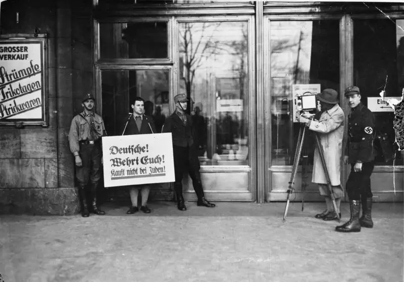 Uniformed men stand outside the entrance of a large department store; one has a placard hung round his neck and a man with a film camera stands at the right.