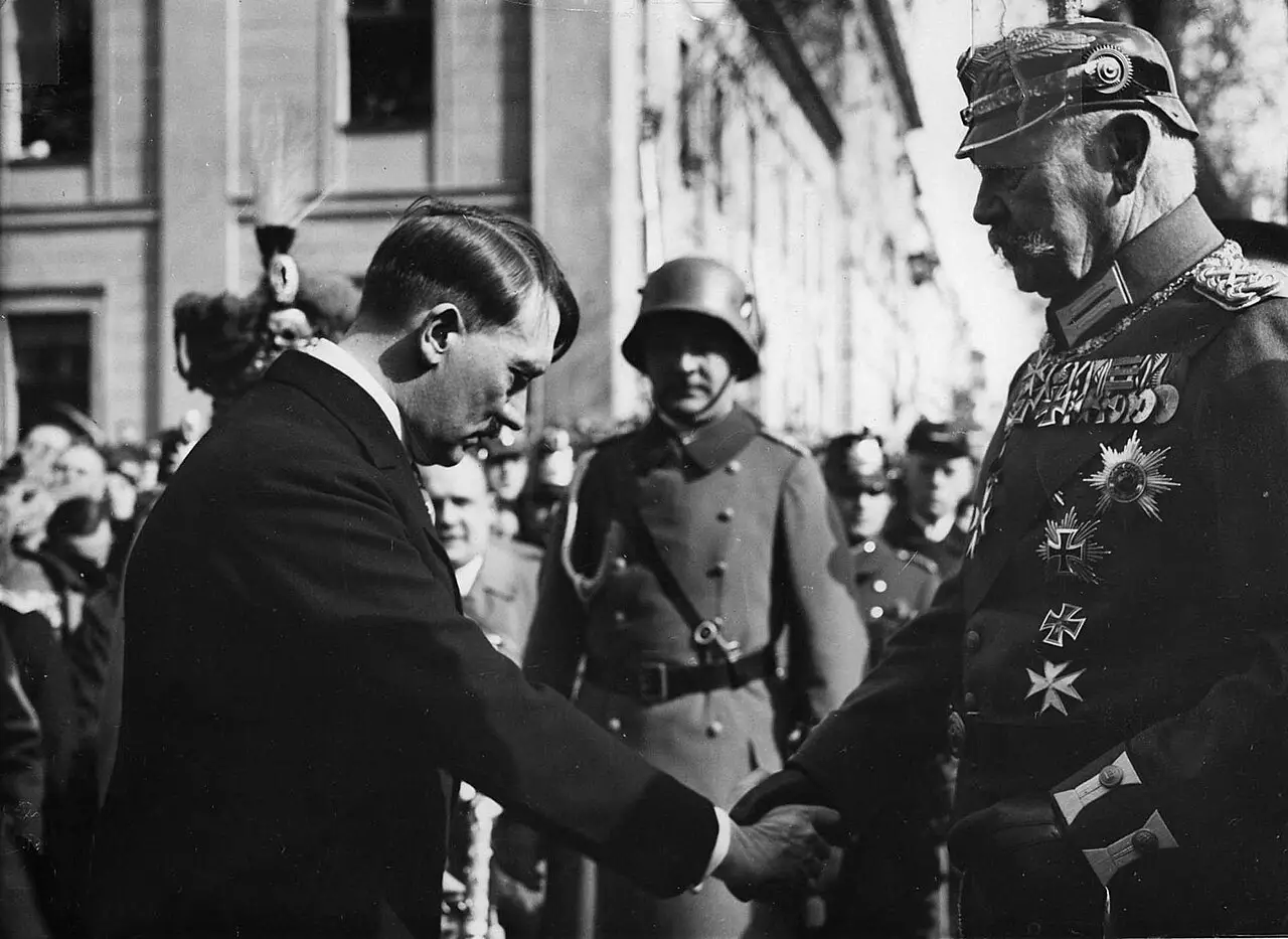 A man in a dark civilian coat bows and shakes hands with an elderly man in a spiked helmet and decorated uniform; a soldier in a steel helmet stands behind.