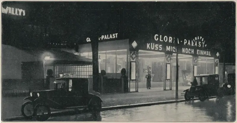 A cinema entrance lit up at night, with the name Gloria-Palast in lights above glass doors and two motor cars at the kerb.
