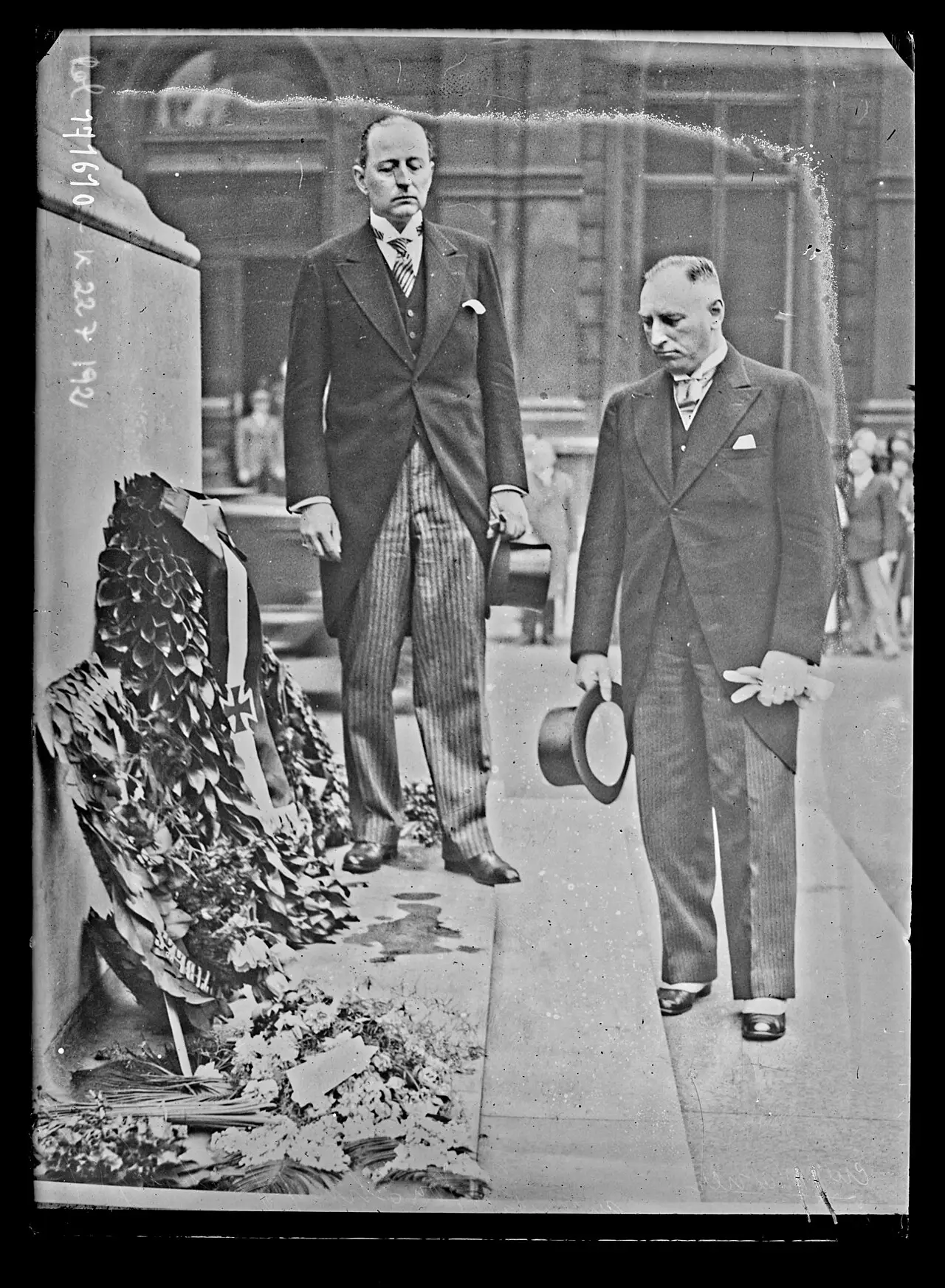 Two men in morning coats and striped trousers stand beside wreaths at the foot of a stone memorial; one holds a top hat.