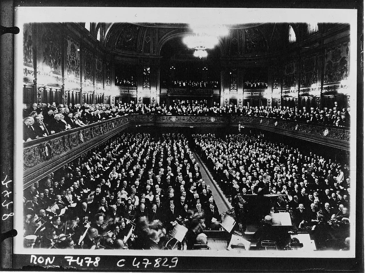A large concert hall seen from above, with an orchestra on the platform in the foreground and every seat in the stalls and galleries filled