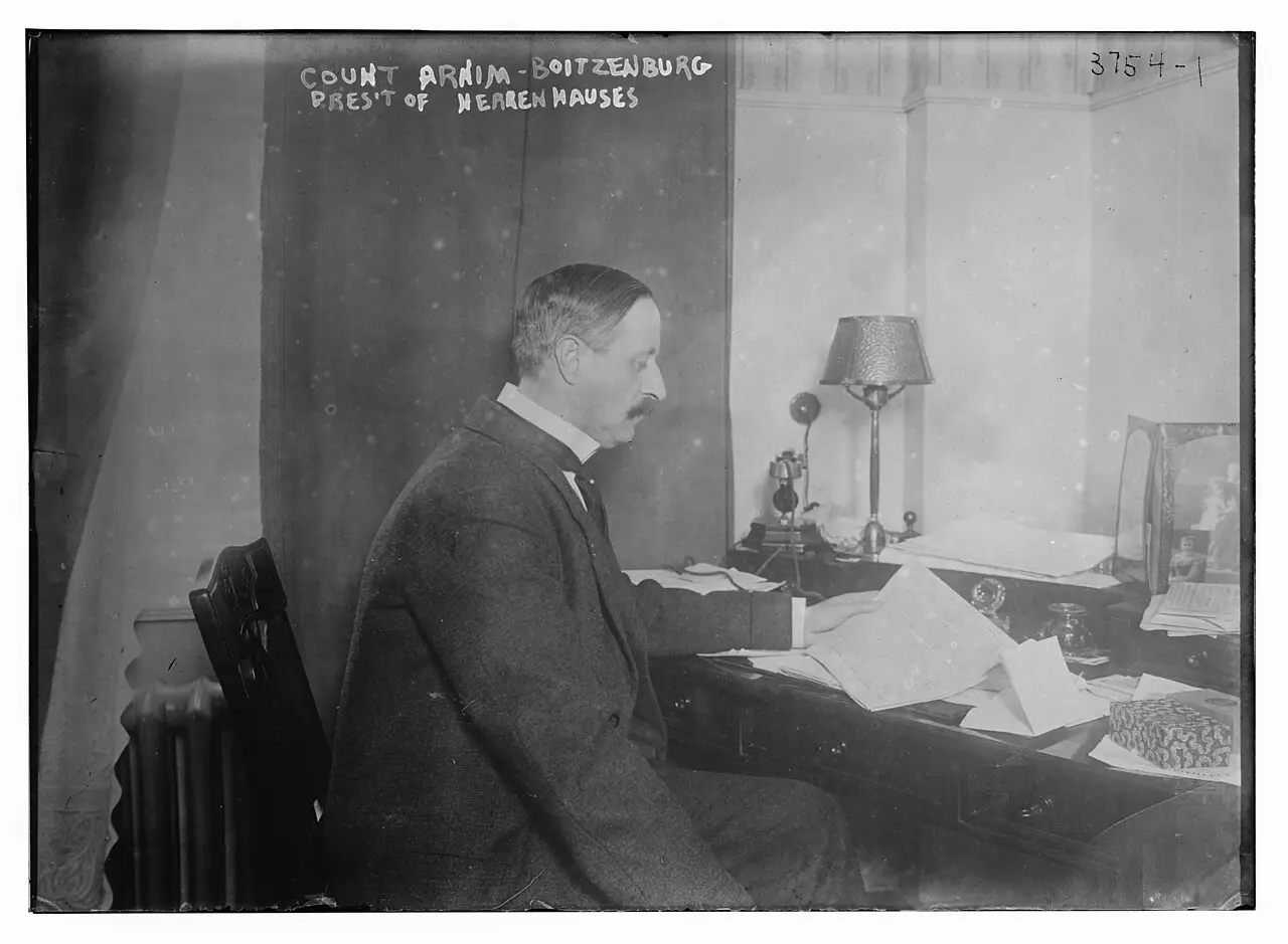 A moustached man in a dark suit sits at a cluttered desk with a lamp and a telephone.