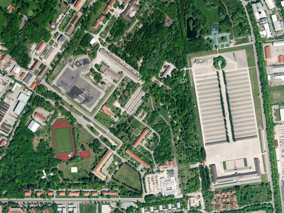 Vertical aerial photograph showing a large rectangular memorial site with rows of barrack foundations beside wooded grounds and buildings.