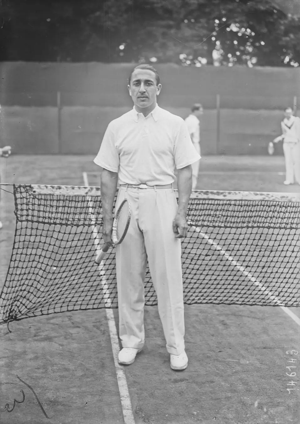 A man in white tennis clothes stands in front of a net holding a racket.