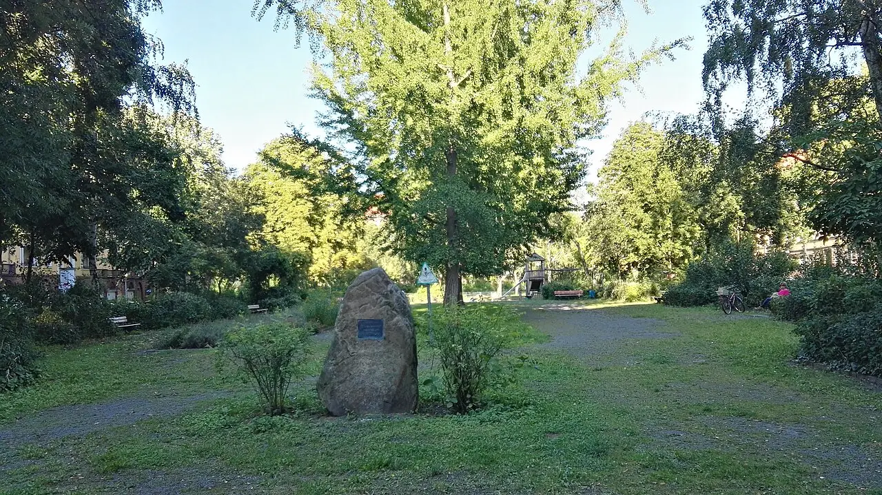 A boulder with a small bronze plaque on a lawn in a tree-lined square.