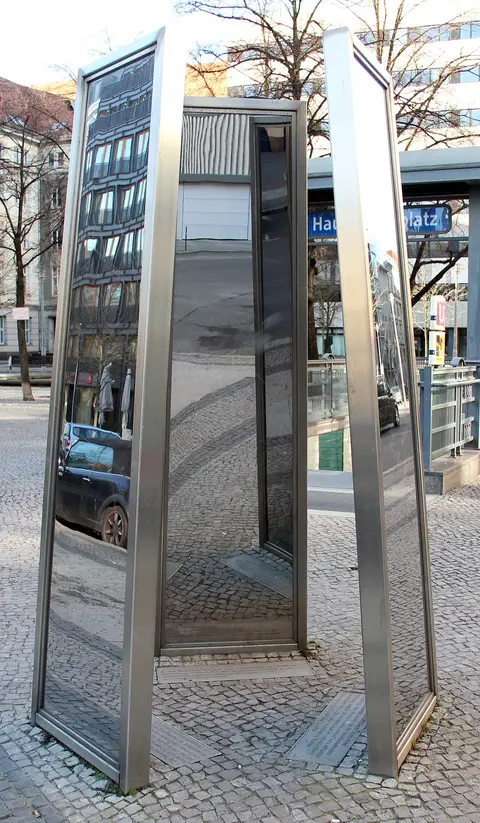 Three tall, narrow mirrored steel panels stand on cobblestones, reflecting the surrounding buildings and parked cars; a U-Bahn station sign is visible behind.