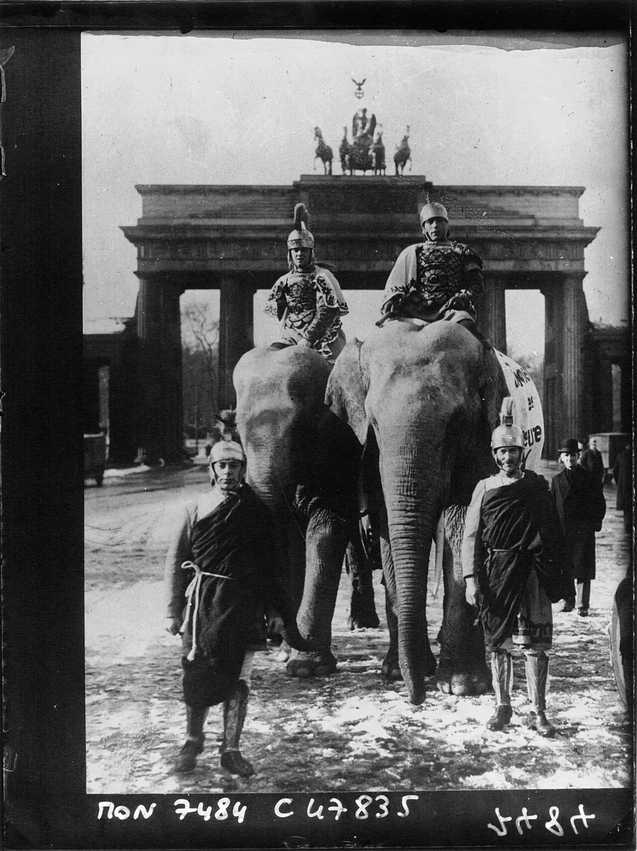 Two elephants ridden by men in Roman helmets and armour cross a snowy square in front of the Brandenburg Gate, with two costumed men on foot