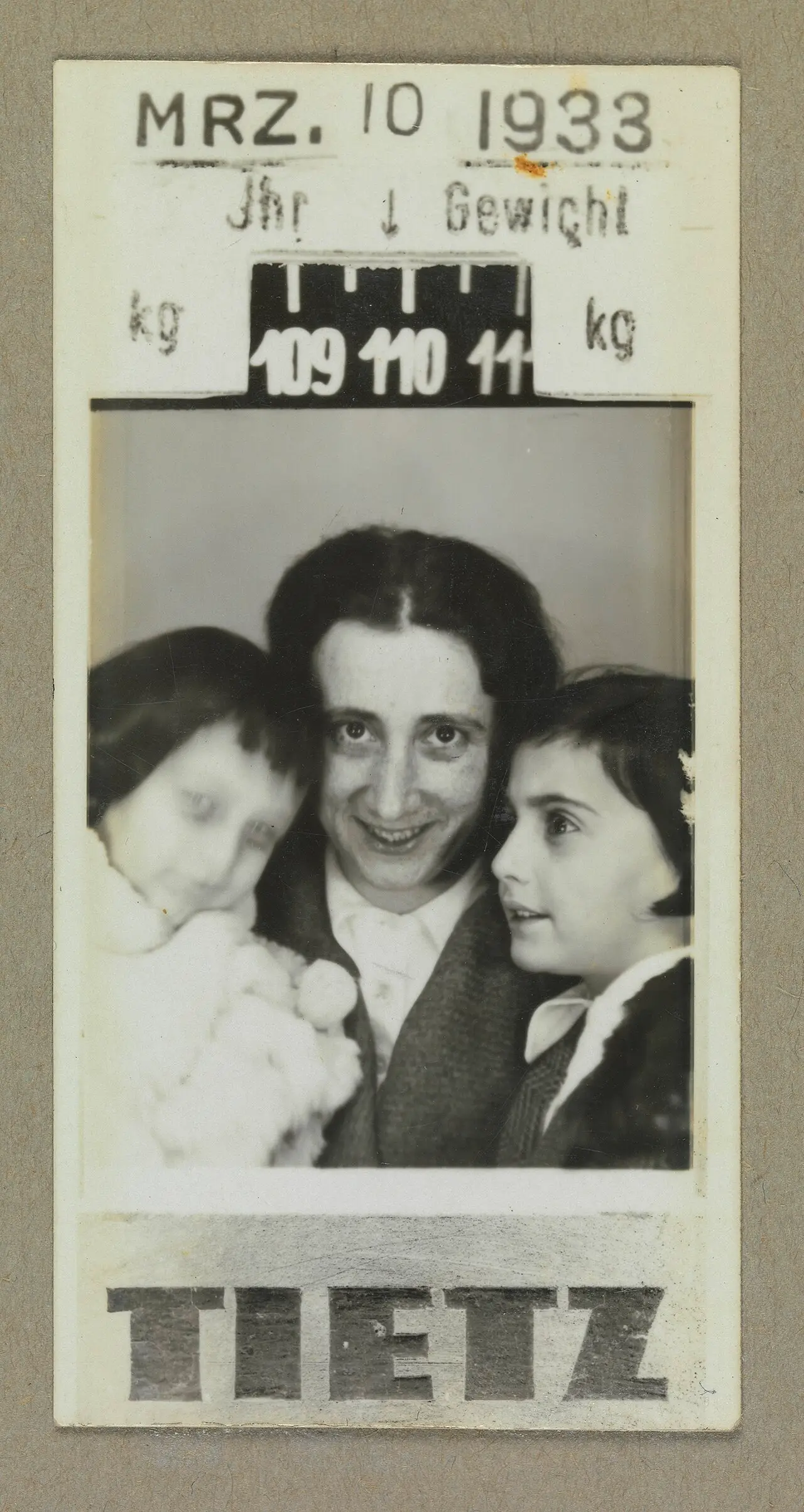 A photo-booth print stamped 'MRZ. 10 1933' above a weighing-scale reading and below it the store name TIETZ, showing a smiling woman with dark hair between two young girls, one blurred and wearing a white fluffy coat.