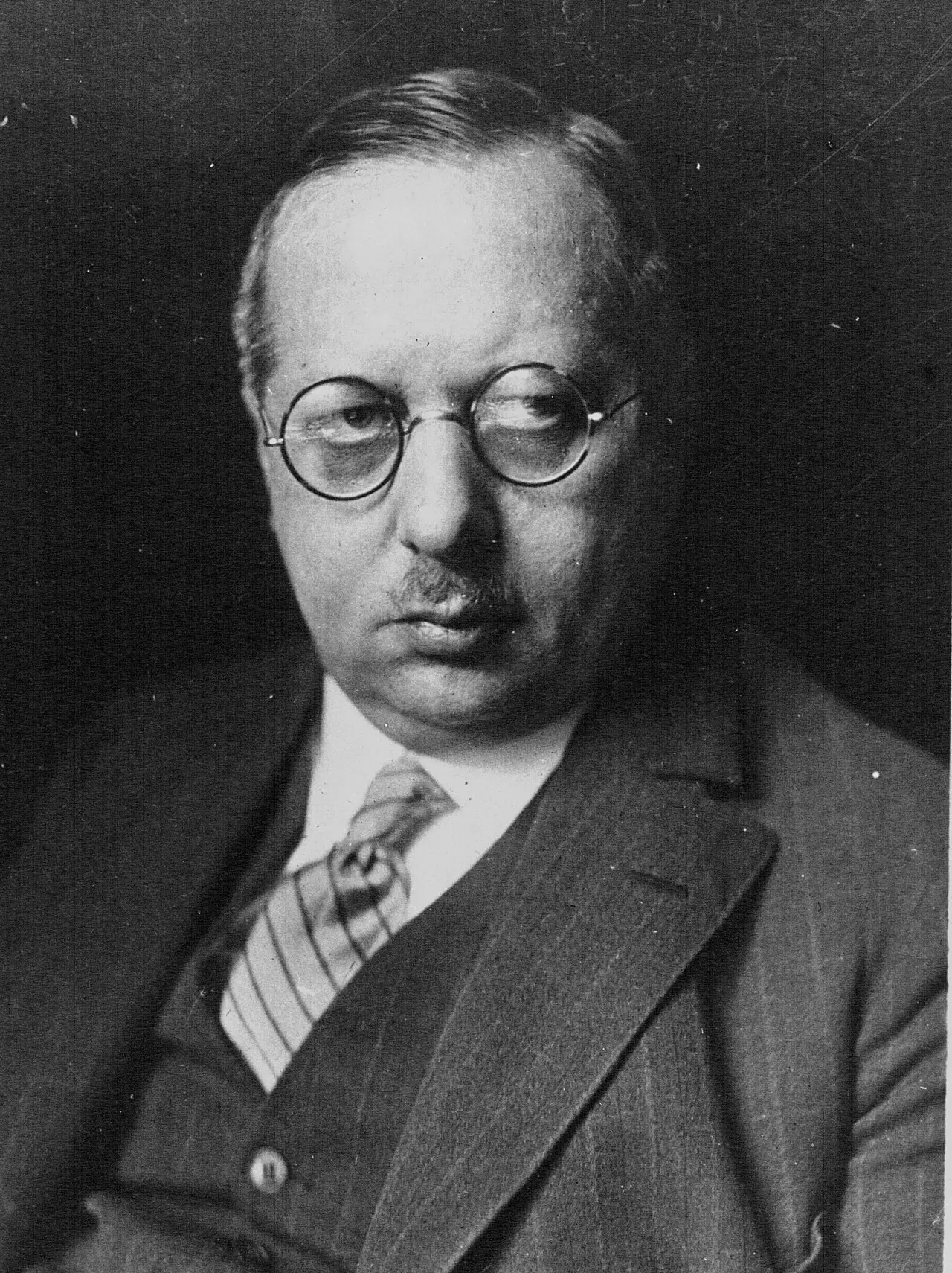 Portrait of a man with round spectacles and a small moustache, wearing a pinstriped suit and striped tie.