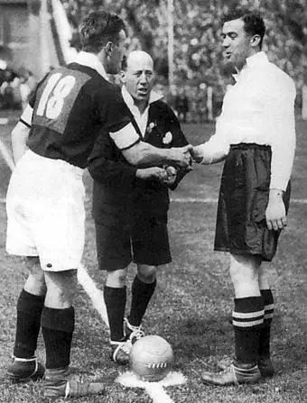 Two footballers shake hands in front of a man in a dark referee's jacket, with a ball at their feet and a packed stand behind