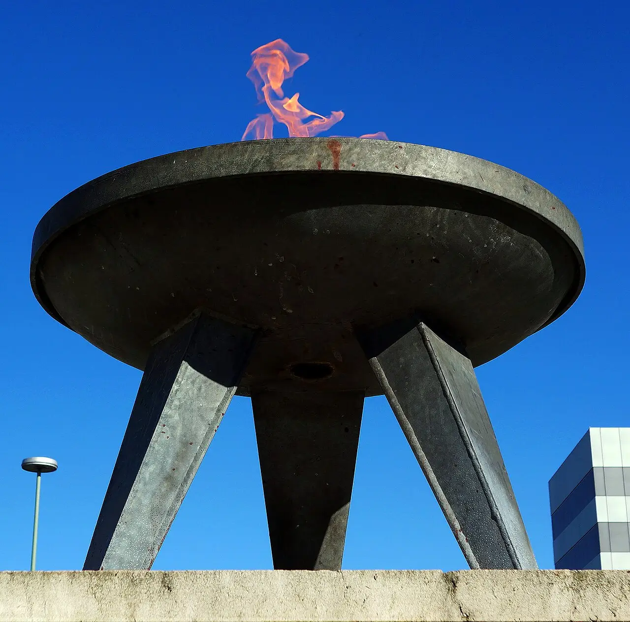 A stone block with an iron bowl holding a flame, on a paved square with trees behind.