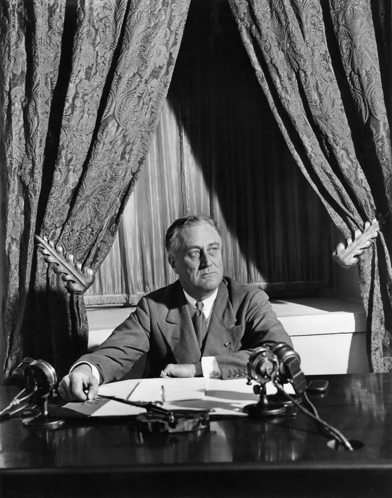 A man in a suit sits at a desk with several microphones in front of heavy patterned curtains, looking up and to the side.