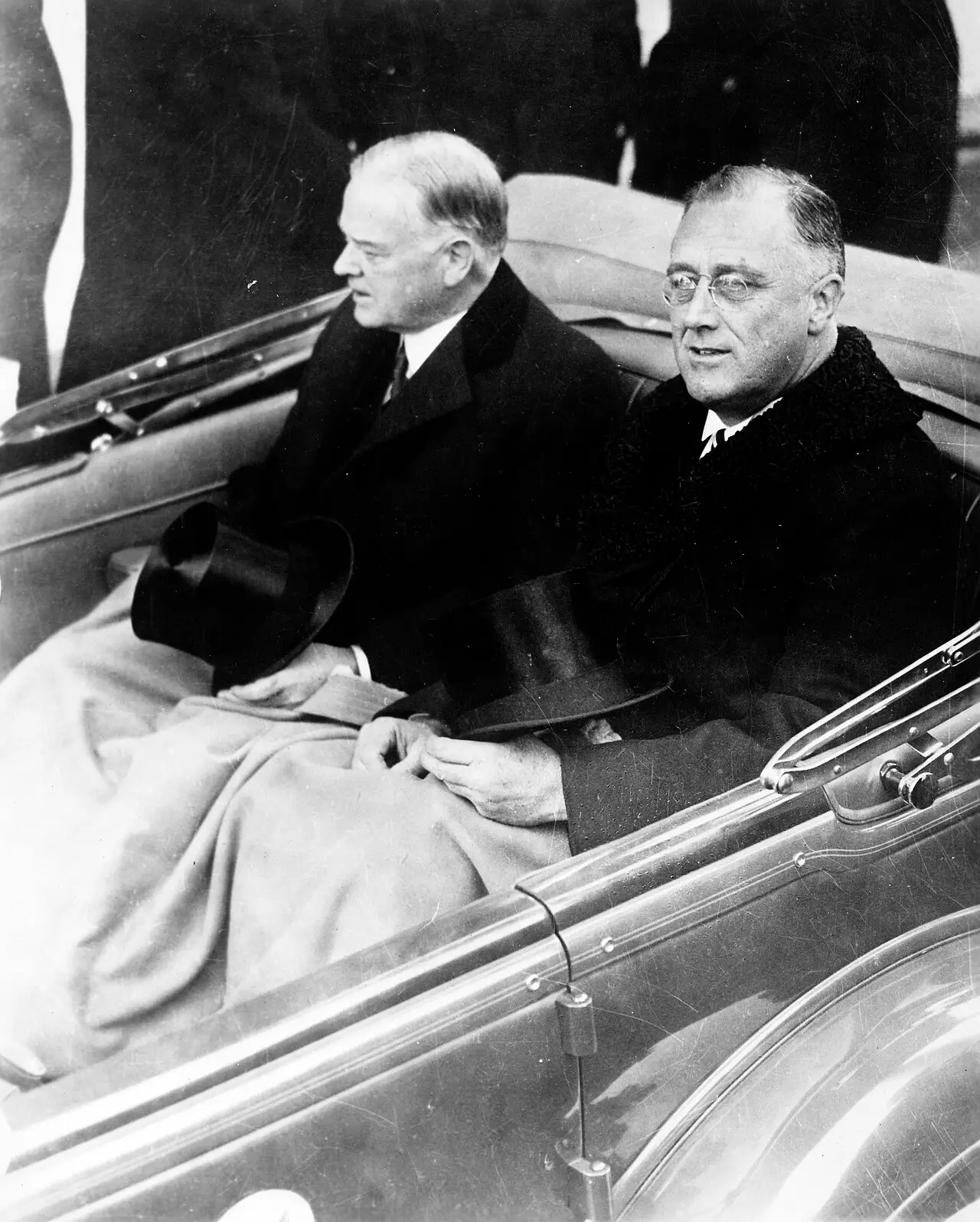 Two men in dark overcoats with top hats on their laps sit under a rug in the back of an open car; the man on the right wears pince-nez.