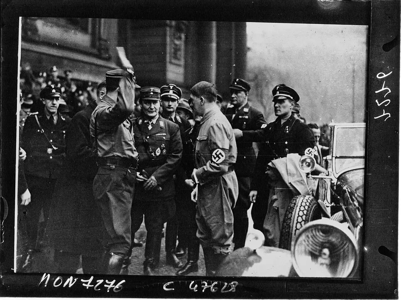Men in party uniforms and peaked caps gather beside an open car outside a stone building; one at the left raises his cap