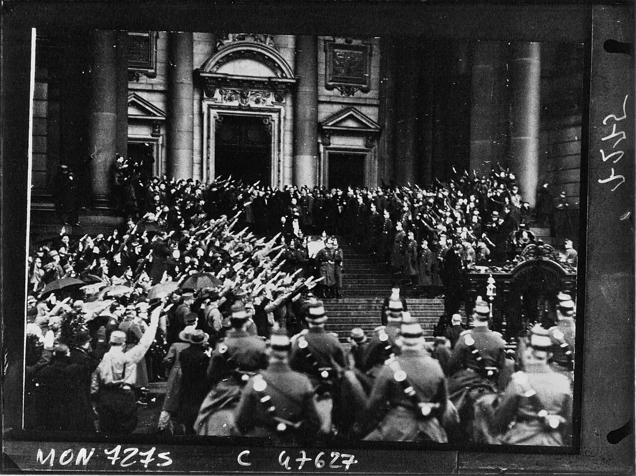 A large crowd with raised arms on and below the steps of a columned church; police in helmets in the foreground