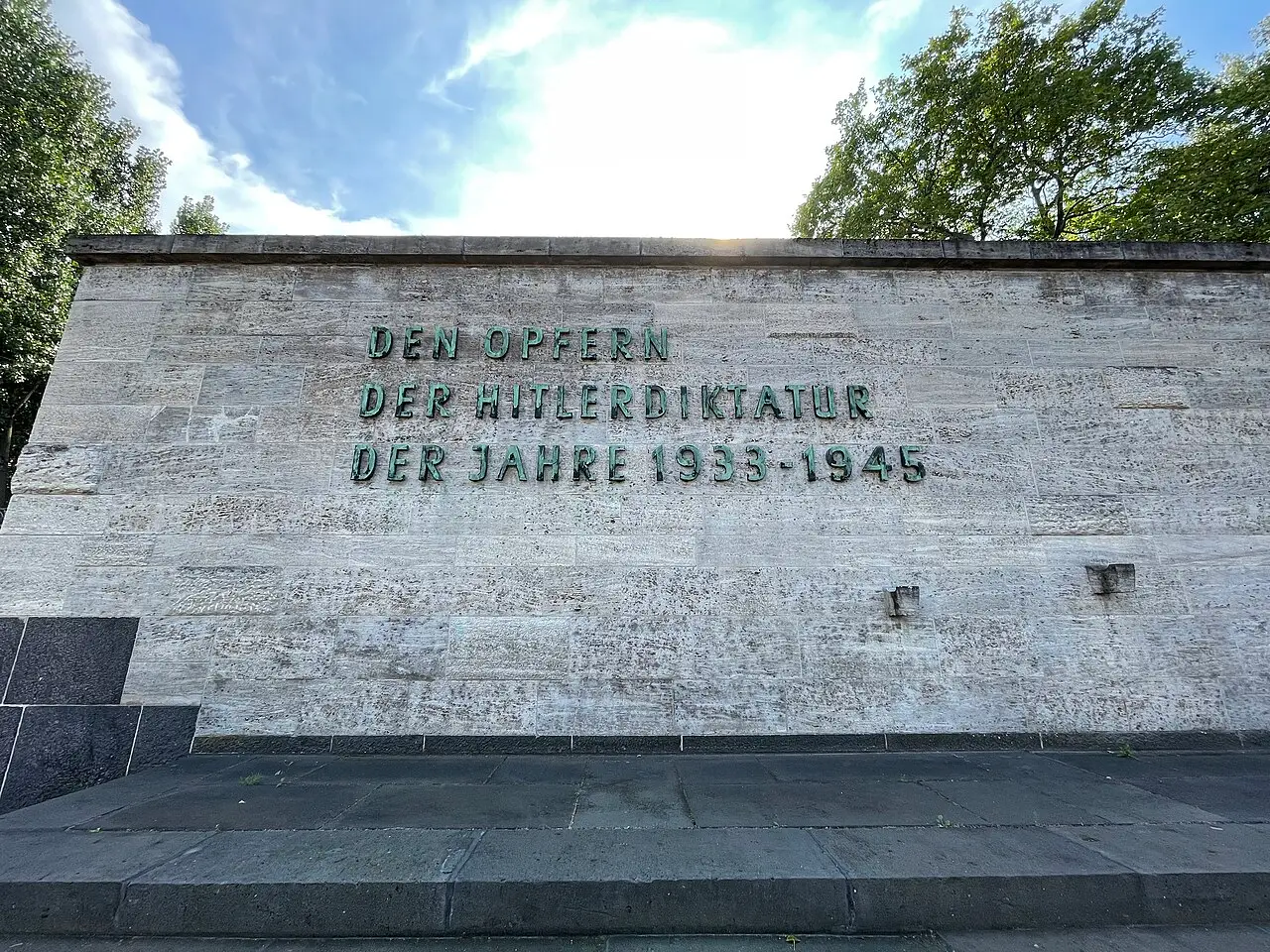 A long wall of pale stone blocks bearing a three-line inscription in green bronze letters, with trees behind it