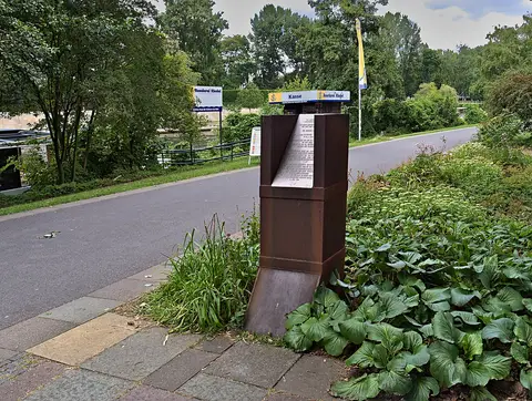 A rust-brown metal stele with an inscribed panel beside a riverside path lined with trees