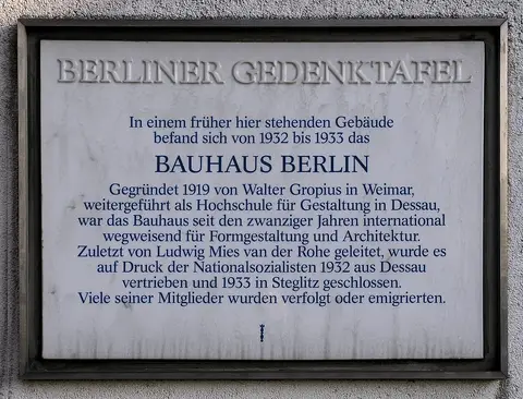 A white porcelain memorial plaque with dark lettering mounted on a wall, headed 'Bauhaus Berlin'.