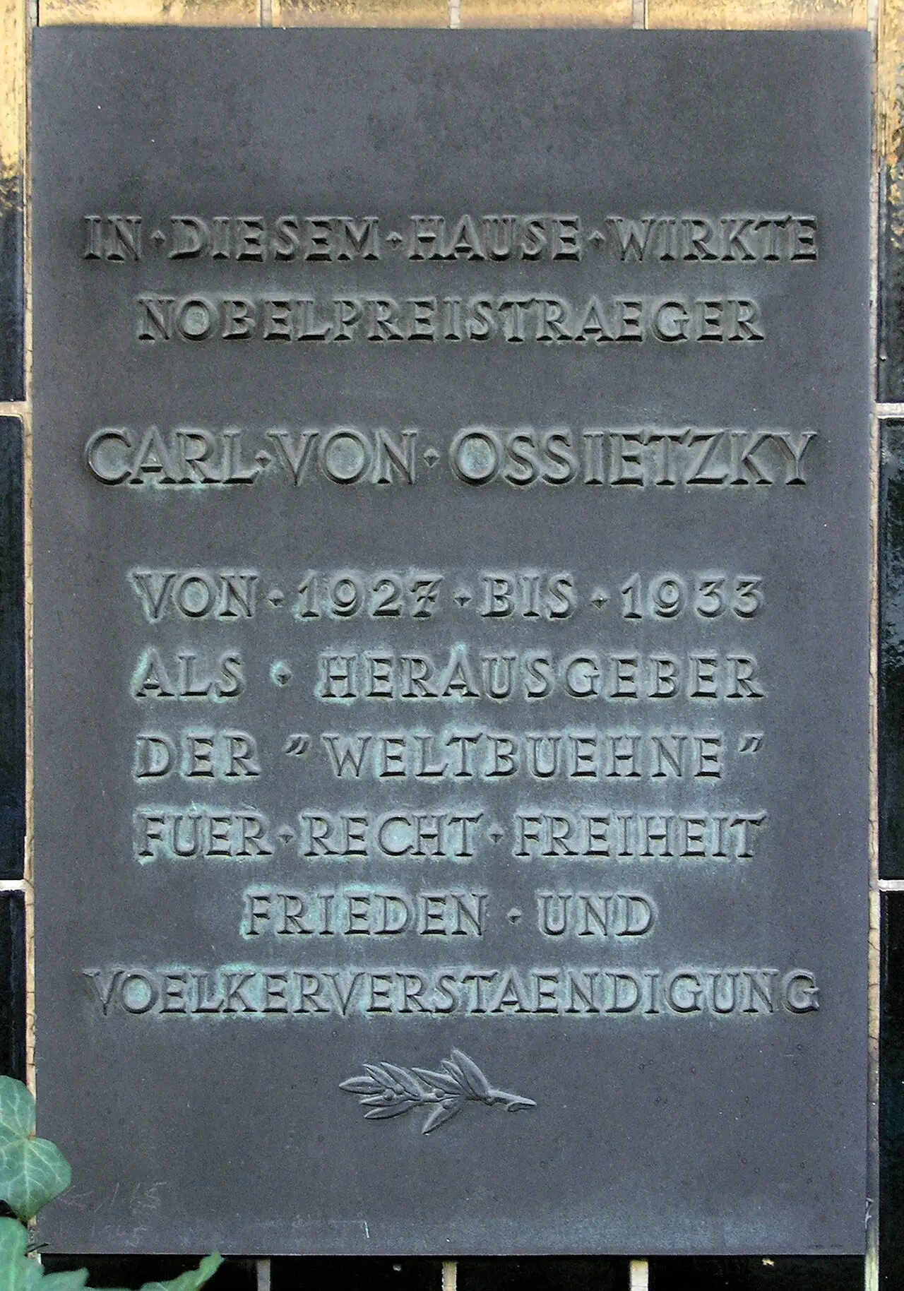A dark bronze plaque with raised capital letters naming Carl von Ossietzky and the Weltbühne, with a laurel sprig at the bottom.