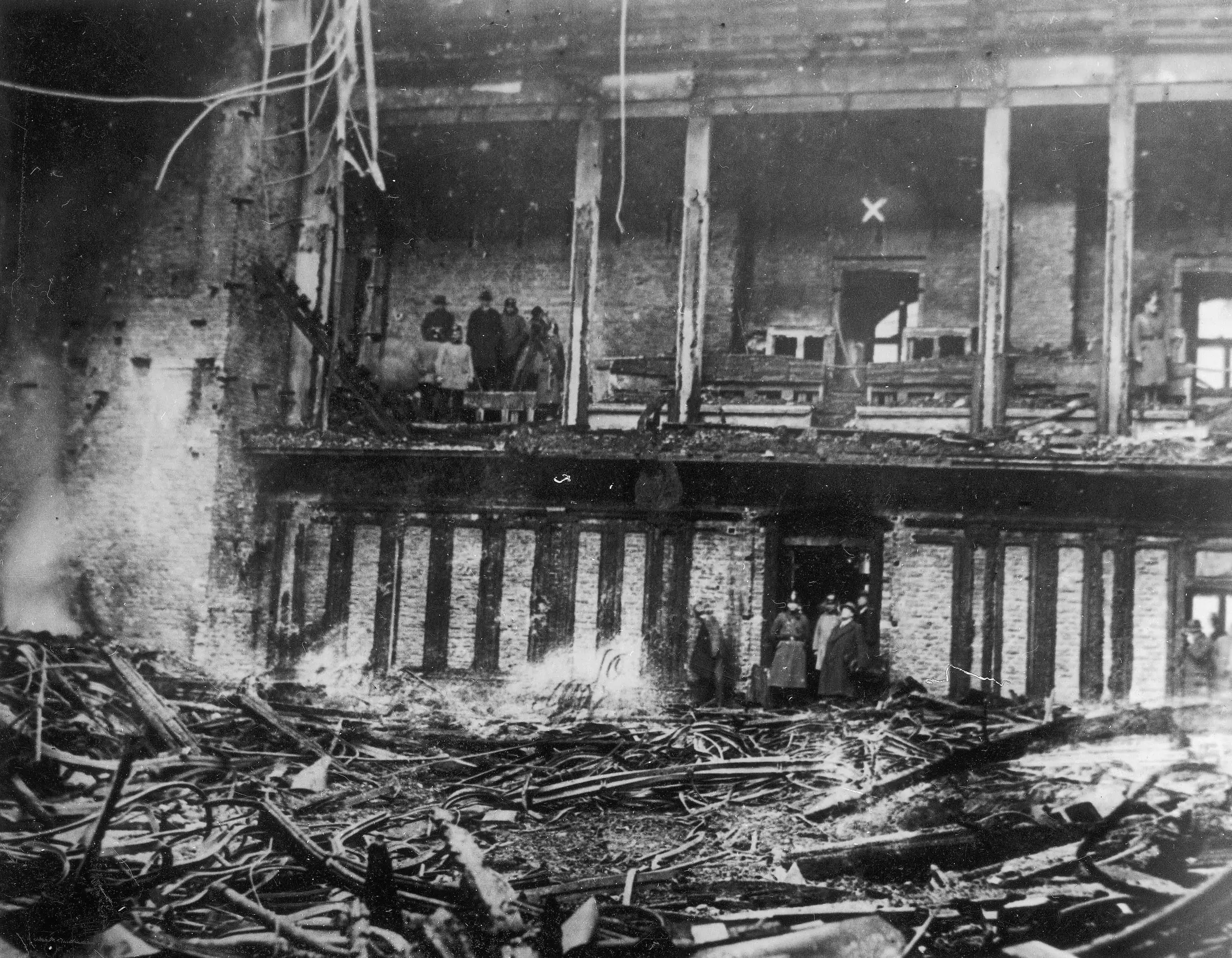 The burnt-out assembly chamber of the Reichstag: charred timber and rubble cover the floor, and a few men stand in the gutted gallery above.