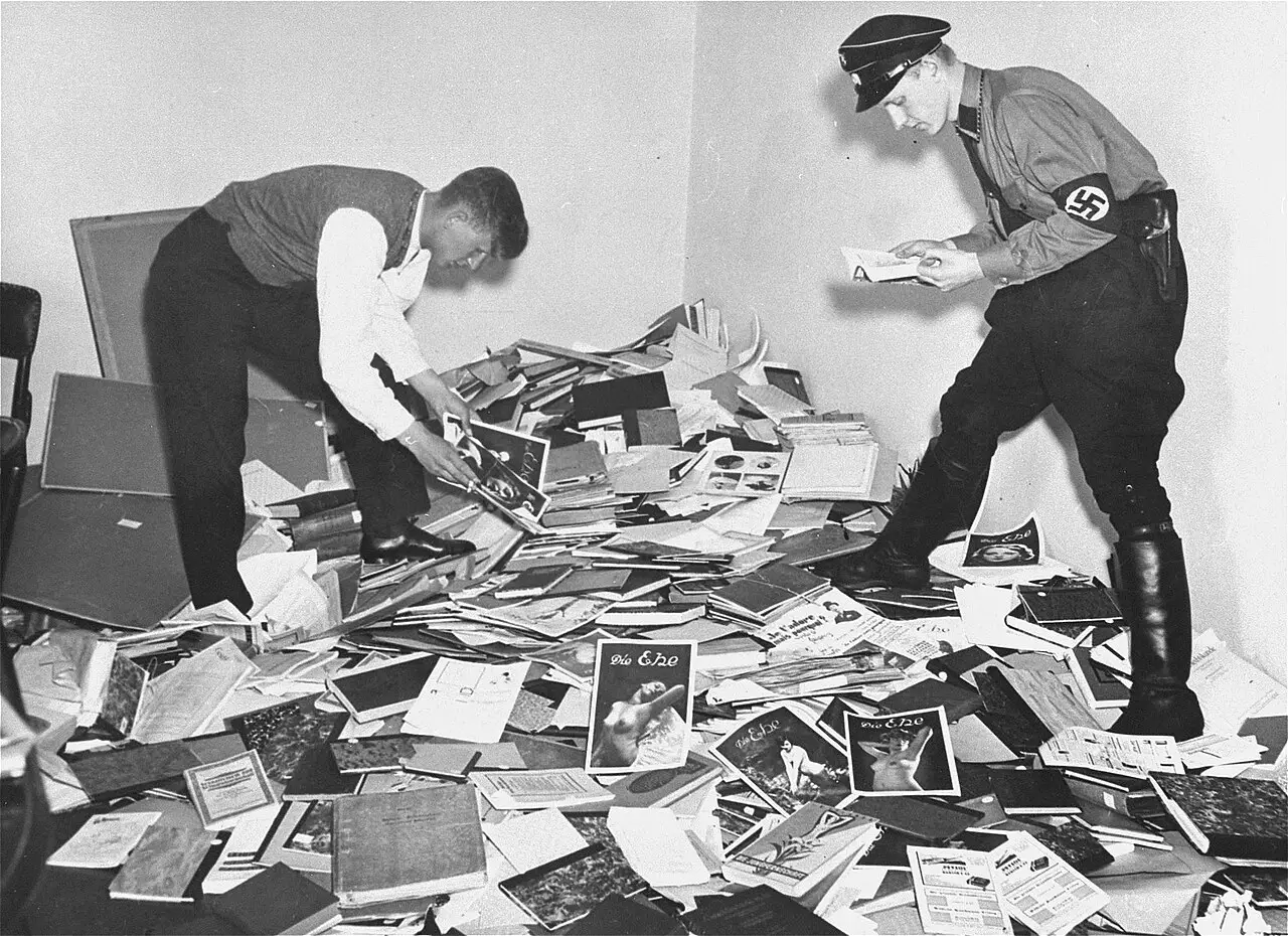 Two young men, one in a black uniform, sorting through a large heap of books, journals and papers