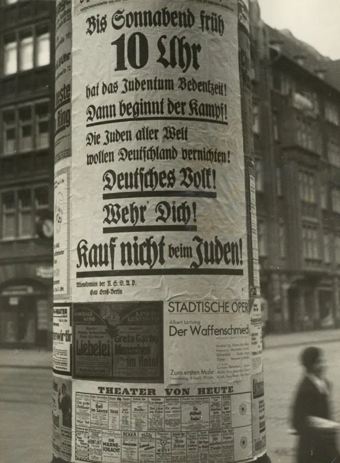 A round advertising pillar in a city street; its upper half is covered by a large poster in heavy Gothic type ending 'Kauf nicht beim Juden!', with theatre and cinema bills pasted beneath.