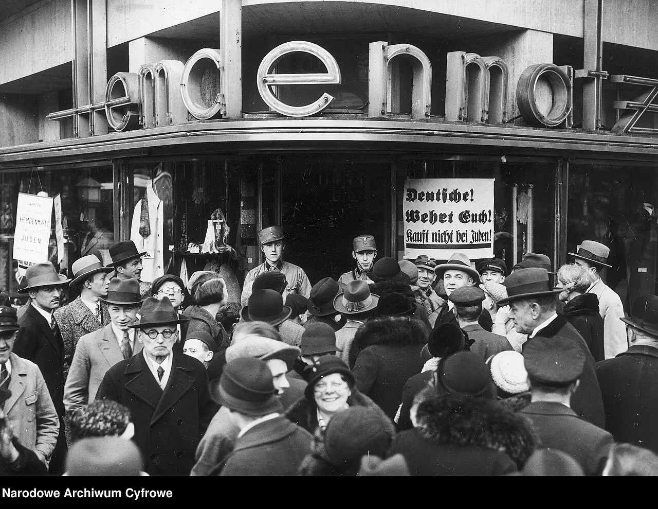 A dense crowd of men and women in hats fills the pavement in front of a modern shopfront with large letters spelling 'Hemdenmatz'; uniformed men stand in the entrance next to a boycott placard.