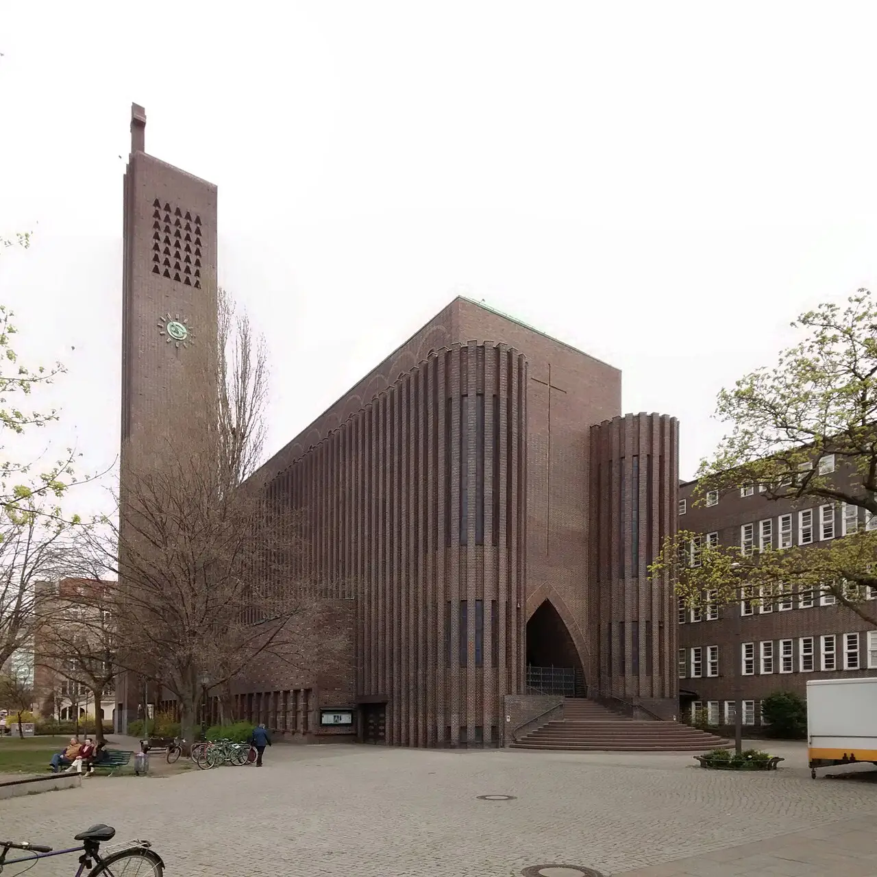 A tall brick church with ribbed vertical piers, a pointed-arch entrance and a slender bell tower, on a paved square.
