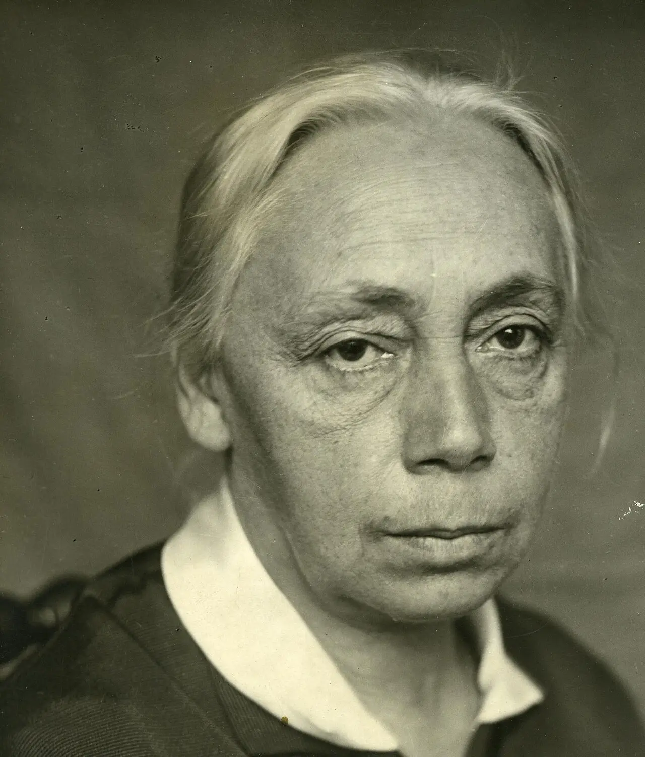 Close portrait of an older woman with grey hair drawn back, a white collar and a dark dress.