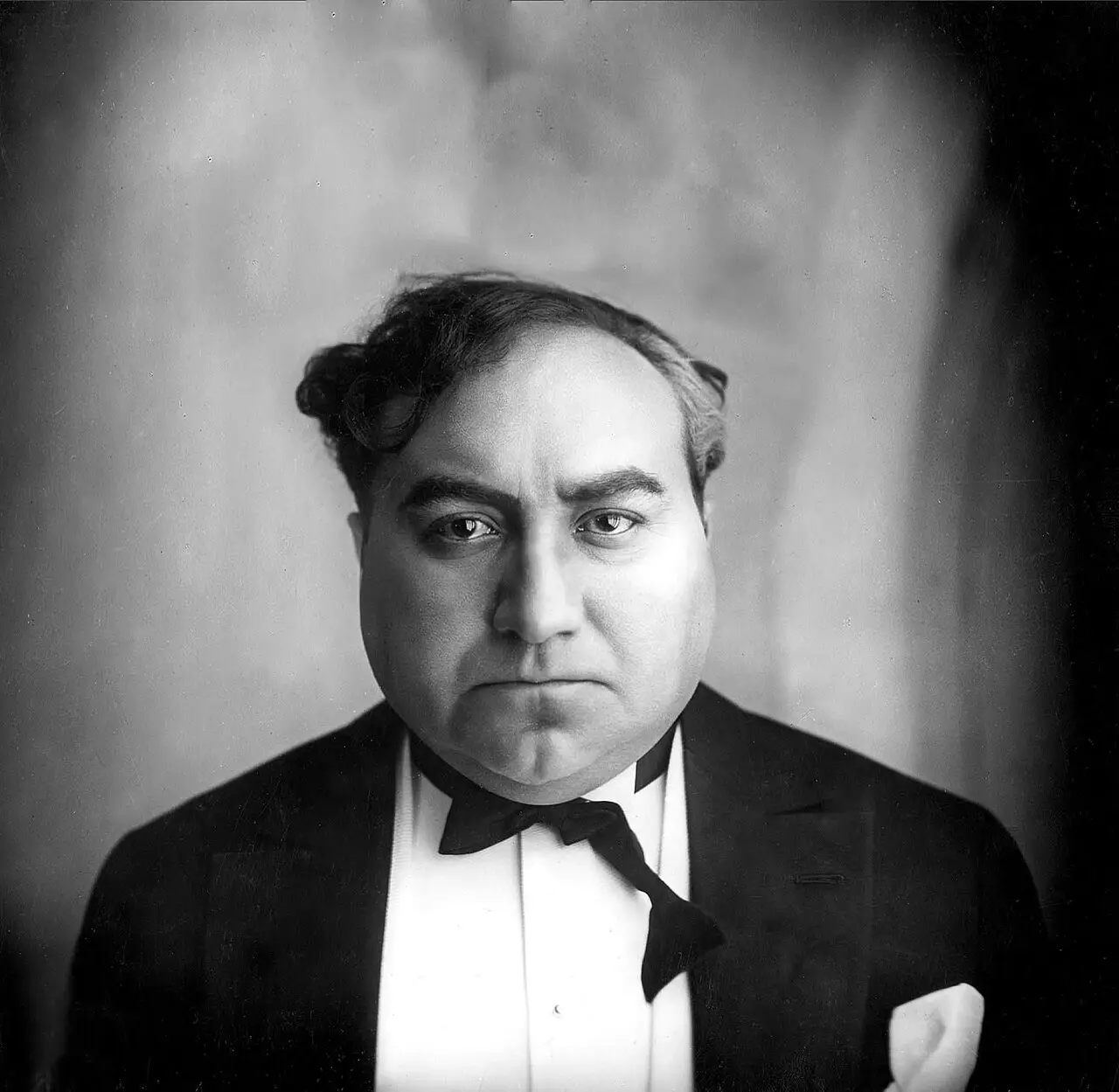 Portrait of a heavy-set man with tousled dark hair, wearing a dinner jacket and bow tie.