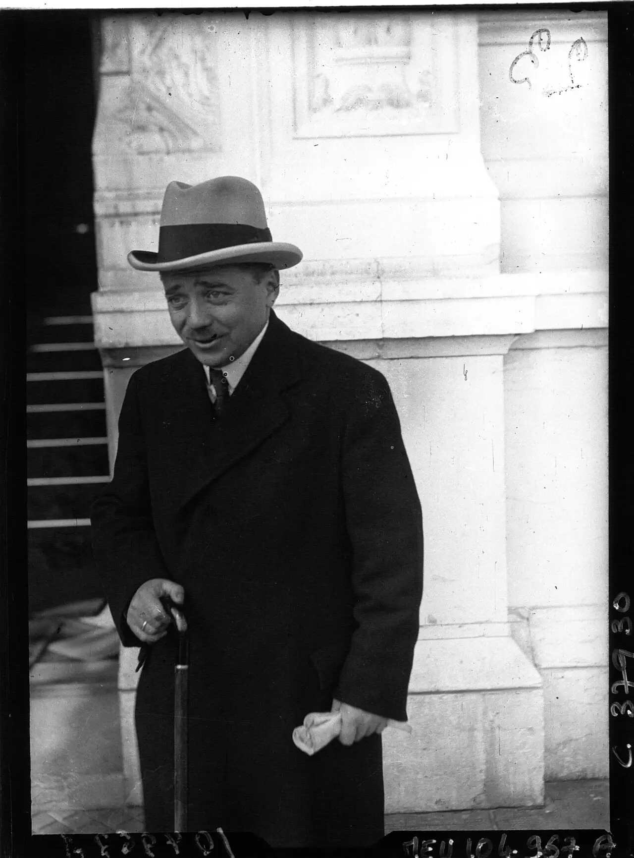 A short man in a dark overcoat and light hat, holding gloves and a walking stick, standing by the stone entrance of a building.