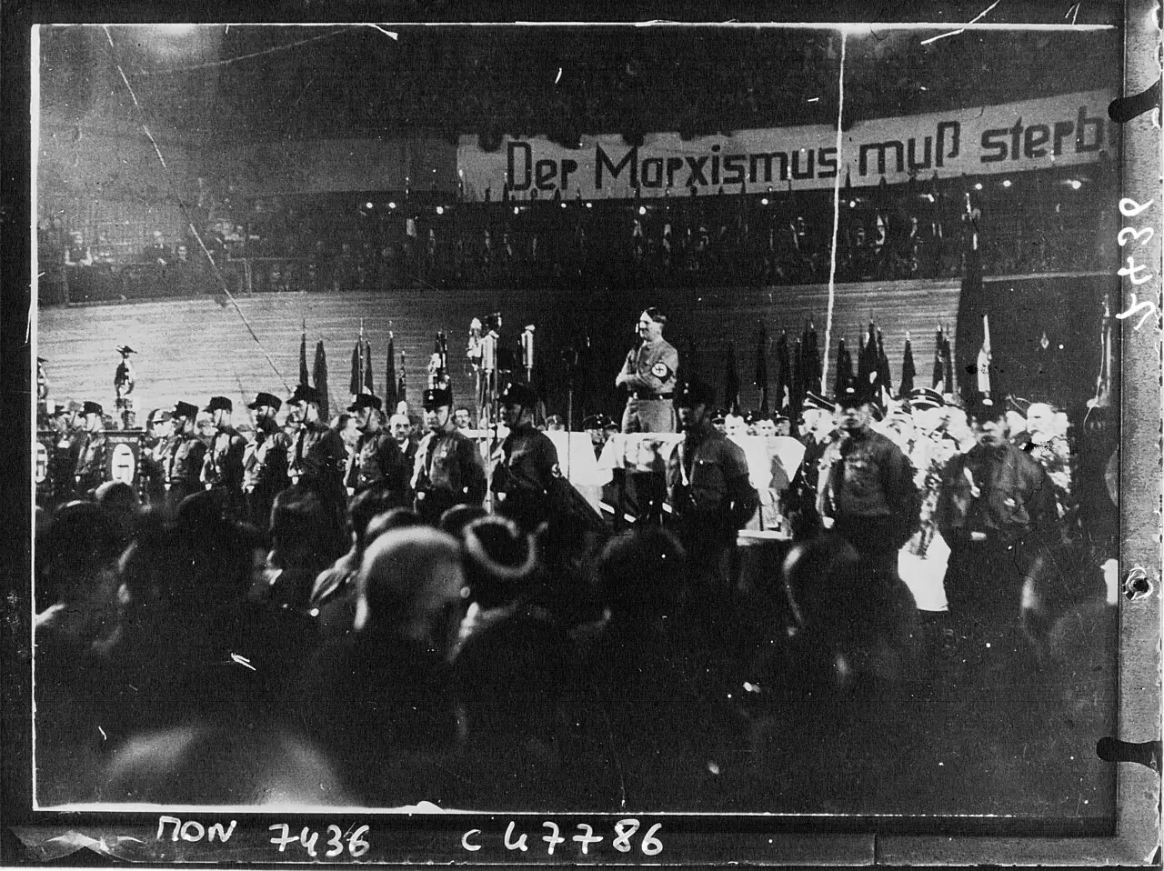 A man at a lectern on a stage in a large hall, flags behind him and uniformed men below