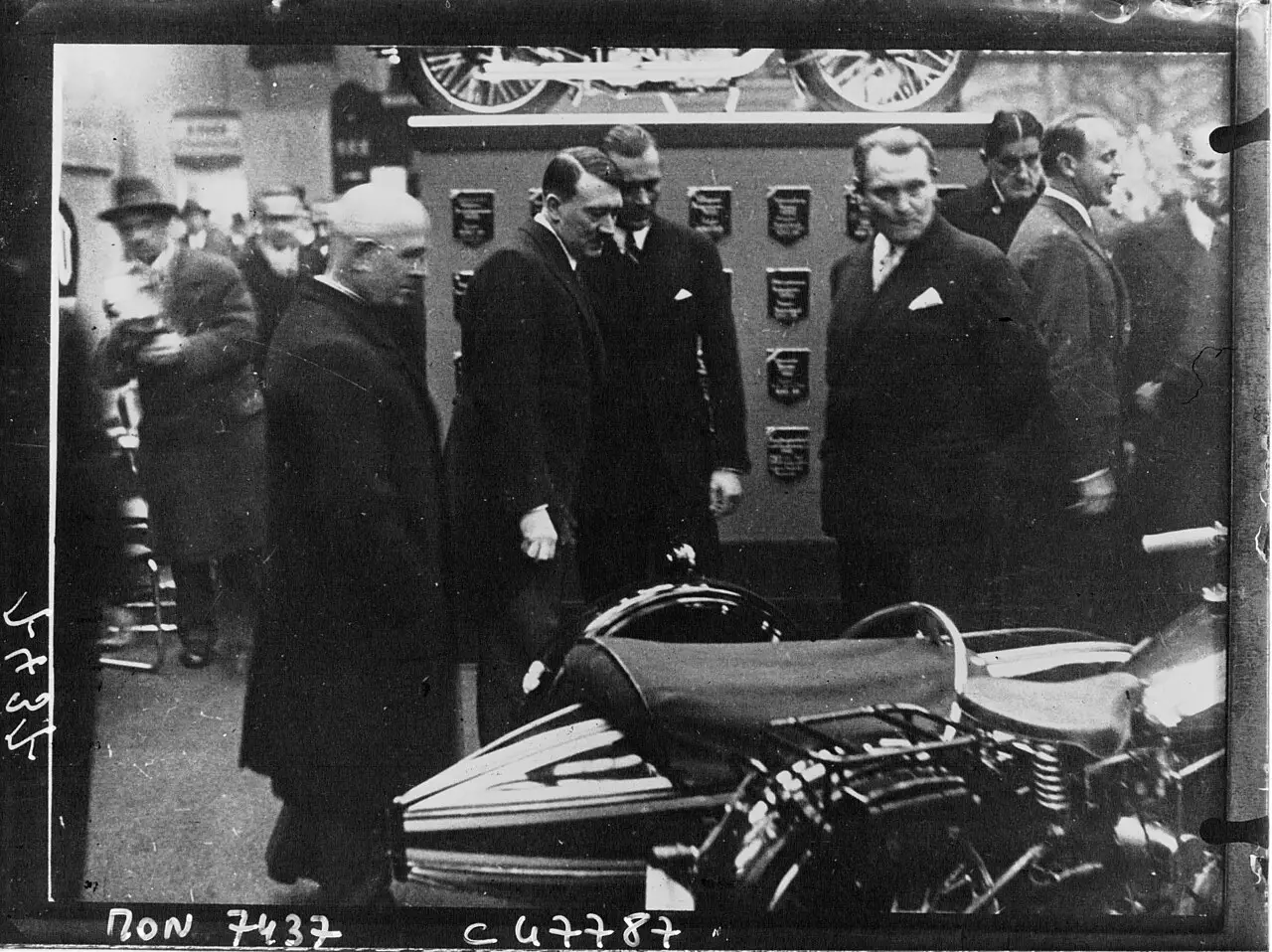 Men in dark overcoats stand beside a motorcycle and sidecar in front of a display board hung with plaques
