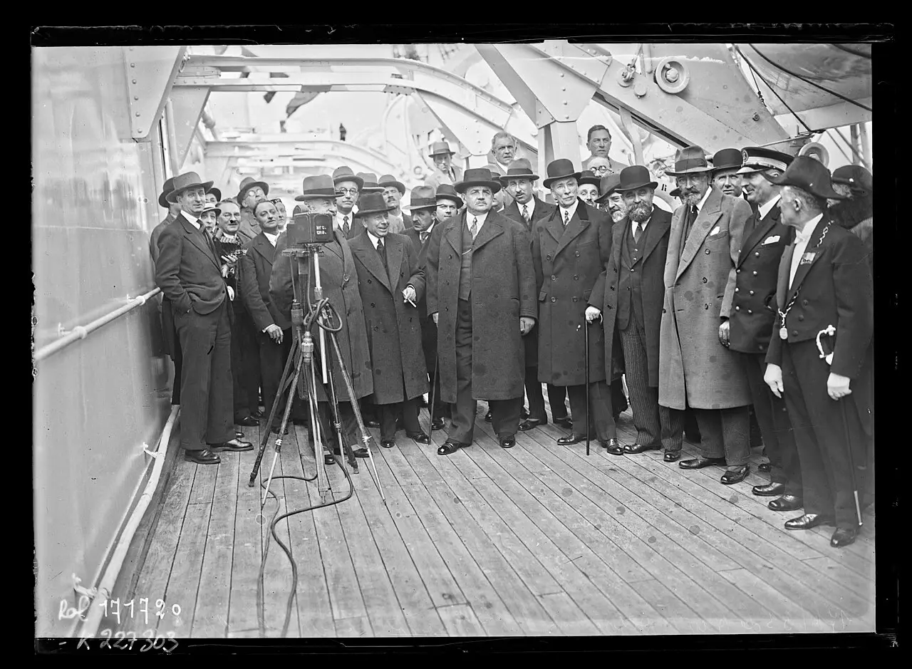A group of men in overcoats and hats stands on a ship's wooden deck beneath steel davits, facing a film camera on a tripod at left.