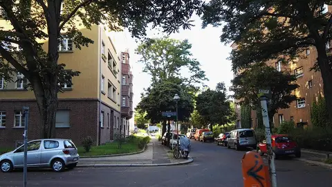 A quiet tree-lined residential street with parked cars between four- and five-storey apartment blocks with rendered upper floors and brick ground floors.