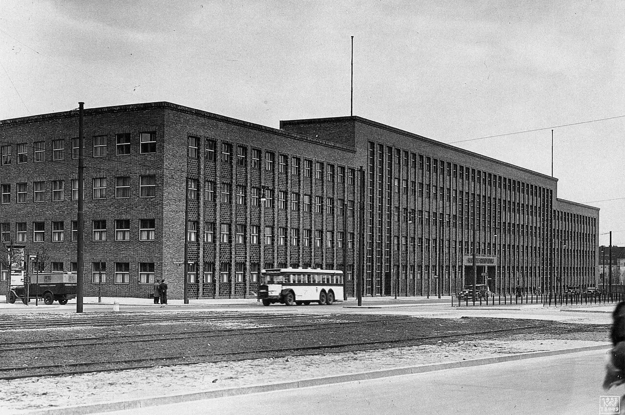 A long, flat-roofed brick office building with rows of tall windows, a double-decker bus passing on the wide street in front.
