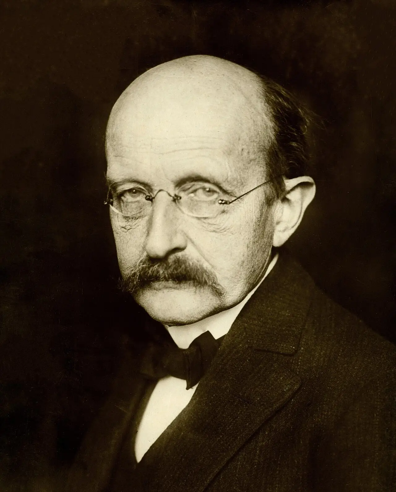 Head-and-shoulders portrait of a bald man with a moustache and rimless glasses, wearing a bow tie