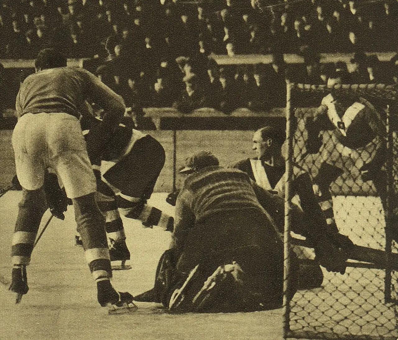 Ice hockey players crowd round a goalkeeper sprawled in front of the net, with spectators packed in the stand behind