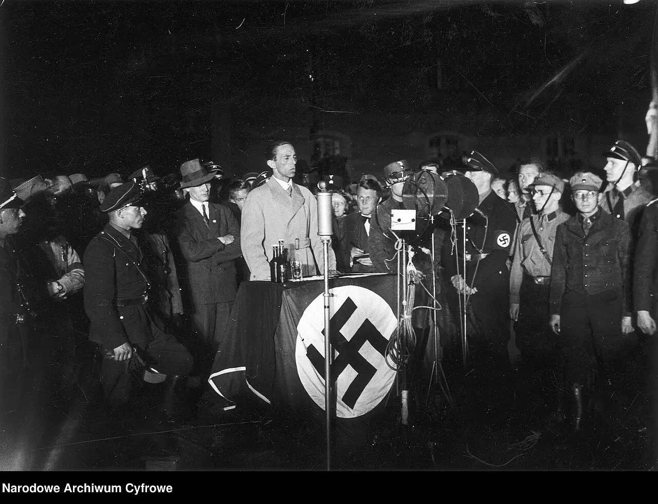 At night, a man in a light coat stands behind a draped lectern with microphones, flanked by men in uniform and in hats