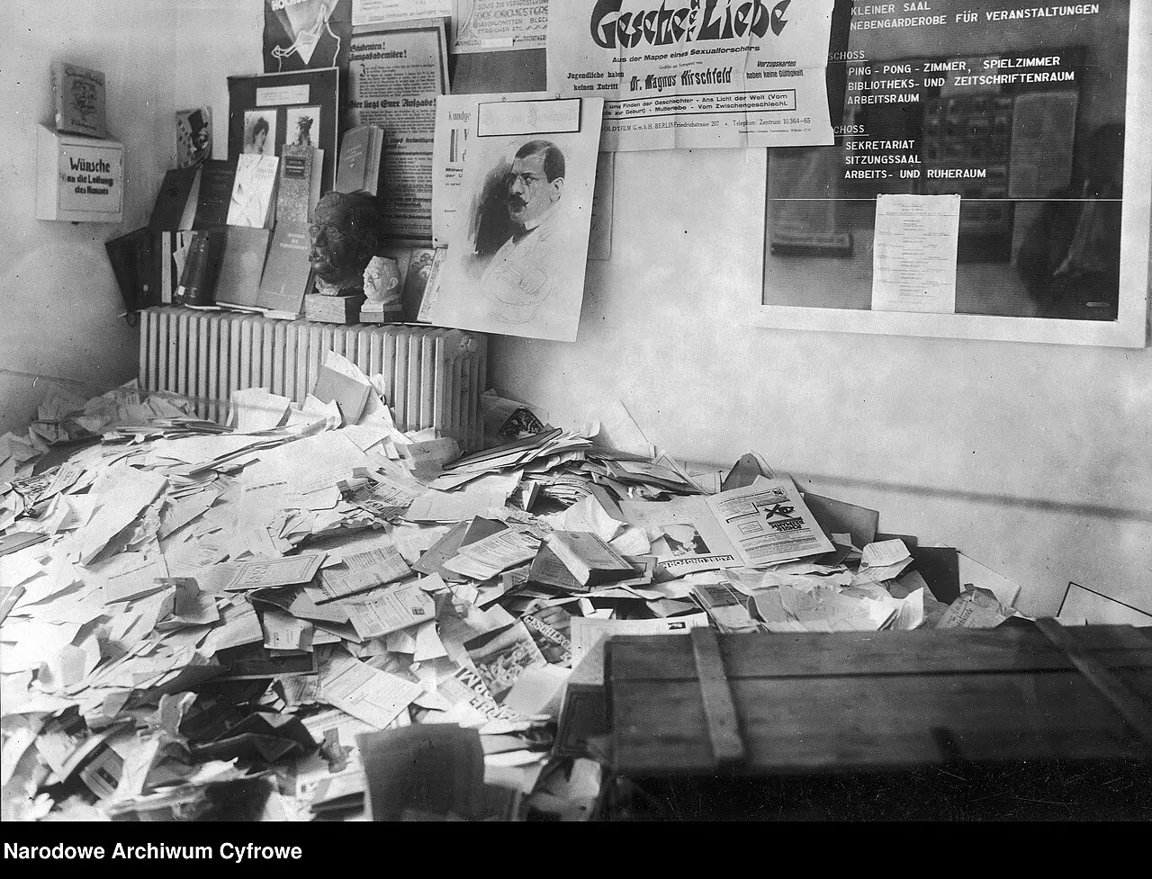 A room with loose papers and pamphlets strewn across the floor beneath a wall of posters, notices and a radiator