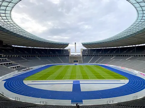 A large oval stadium with a blue running track, a green pitch and grey seating under a curved roof, open at one end towards a tall tower.