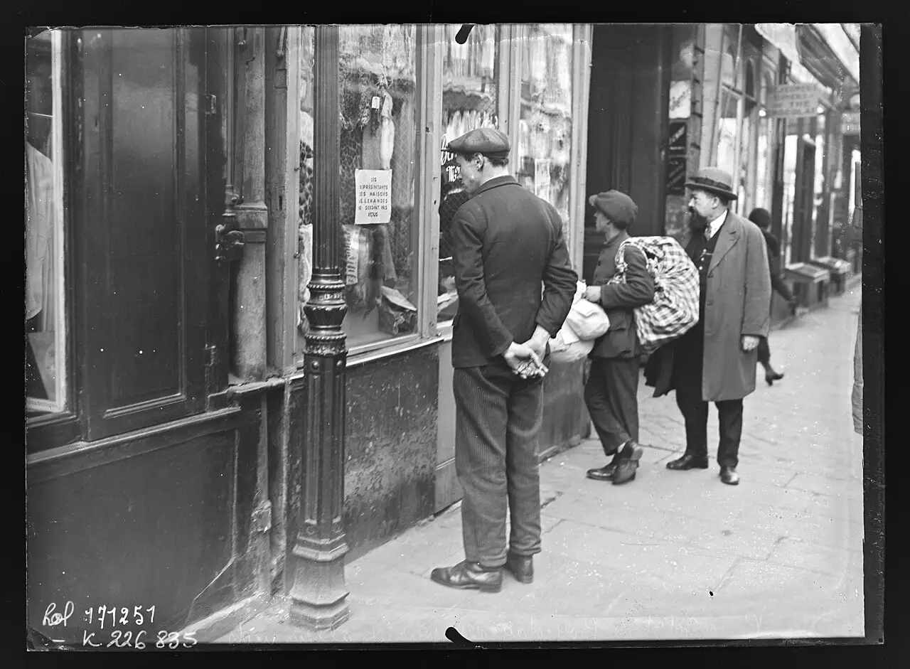 On a Paris pavement a man in a cap stands with his hands behind his back reading a small printed notice in a shop window hung with lace; a boy carrying a large checked bundle and an older bearded man walk past.